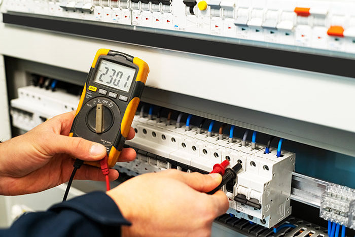 Primo piano sulle mani di un tecnico elettricista che utilizza un multimetro digitale professionale per misurare la tensione (230.1 V) sui moduli di un quadro elettrico industriale. Si notano i cavi colorati e i dispositivi di sicurezza.