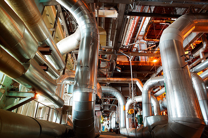 Immagine in primo piano di un labirinto di tubazioni industriali e condotte metalliche in un impianto di processo con illuminazione calda.