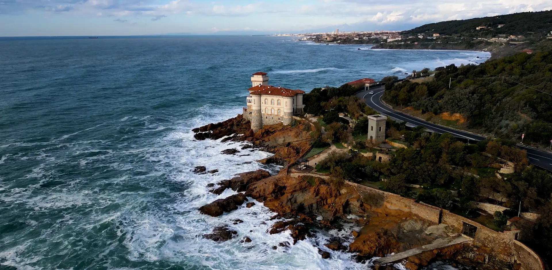 Veduta aerea di Castel Boccale a Livorno arroccato sulla scogliera del Romito; onde del mare mosso si infrangono sugli scogli e strada costiera che attraversa la macchia mediterranea.
