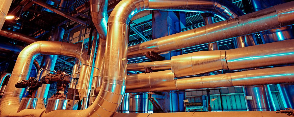 Fotografia industriale di un complesso sistema di tubazioni di acciaio isolato, con valvole, illuminato da un drammatico contrasto di luce blu e arancio all'interno di una centrale elettrica o di un impianto di produzione.